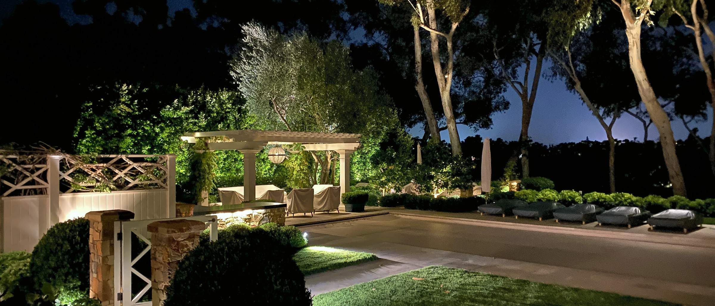 A whole garden at night lit as one system: pergola, lawn, trees and hedges